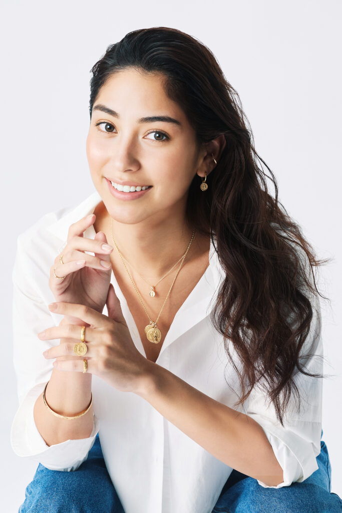 白いシャツを着た黒髪の女性がゴールドのコインネックレスとリングを着用